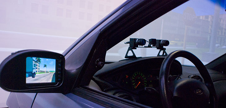 Driver's side of a car with a pair of cameras or projectors mounted on the dashboard and a computer-generated scene in the side mirror
