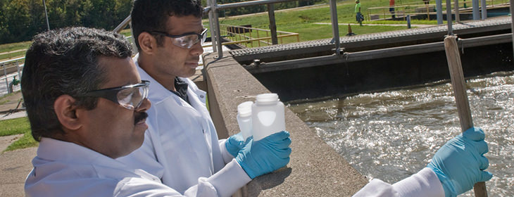 Two people outdoors in lab coats taking water samples from an artificial reservoir