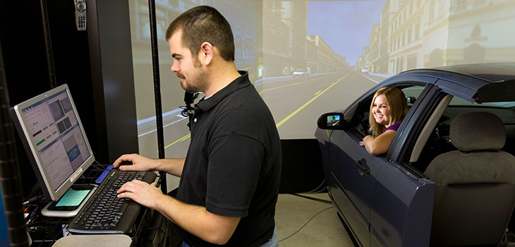 One person sitting in the driver's seat of a car with the part behind the back seats cut away, and a scene of a roadway projected on the wall in front, while another person uses a computer nearby