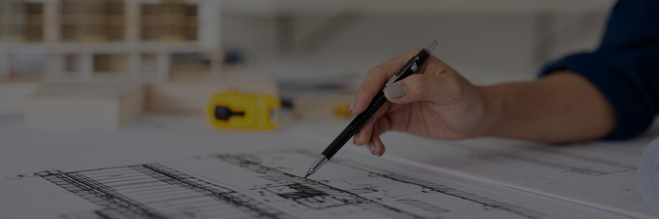 A woman with a pencil and engineering drawings, with a hard hat in the background