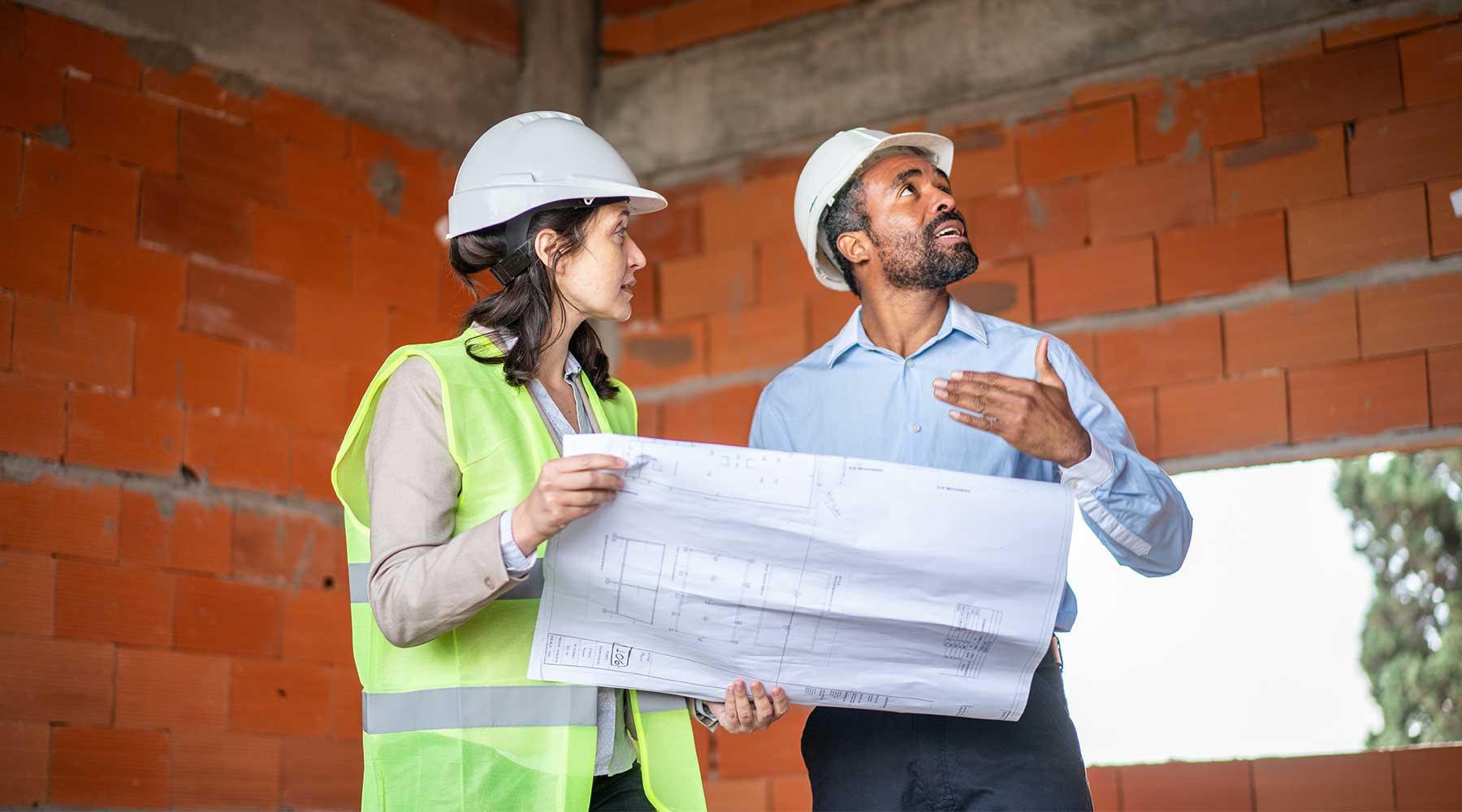 Two engineers holding blueprints next to brick wall