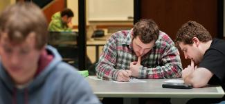 Two students work on a test sitting at the same table, with other working students in the foreground and background