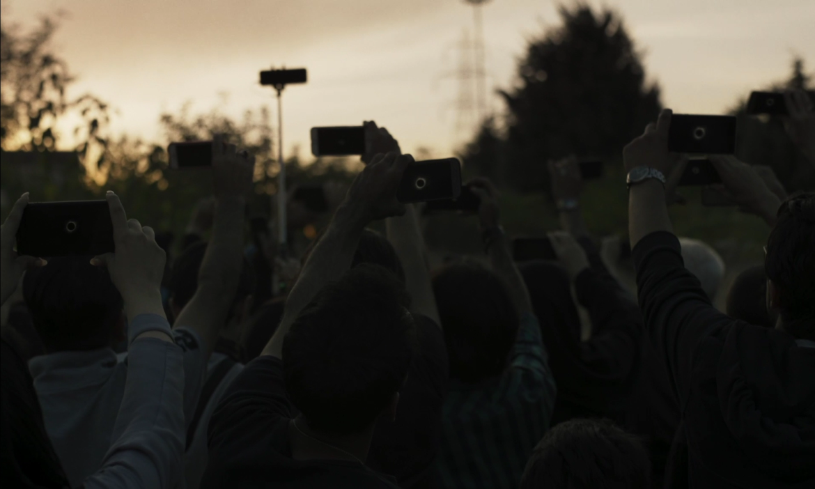 An image from Solar Eclipse, a crowd of people raising up their phones towards the sky.