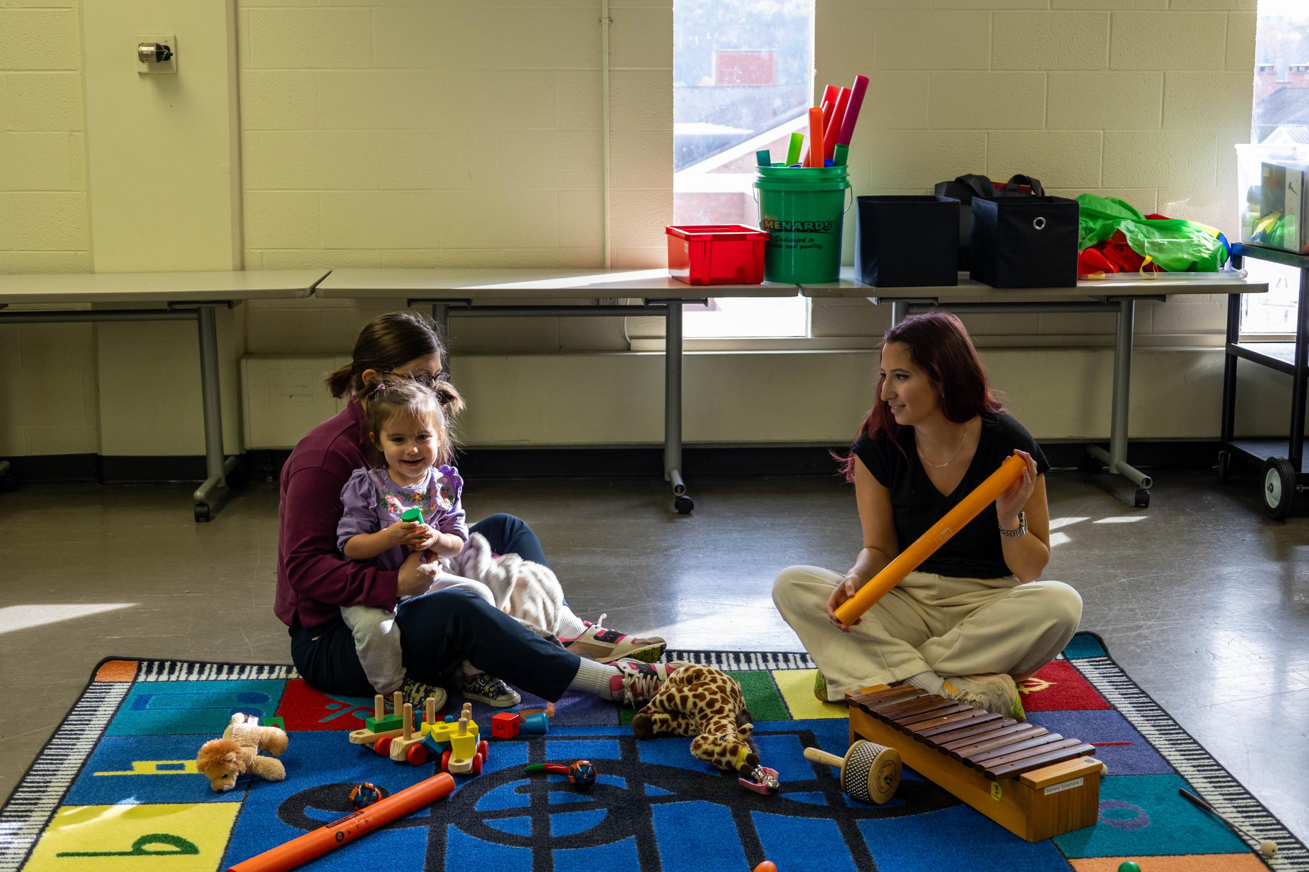 Parent, toddler, and teacher play instruments on a rug.