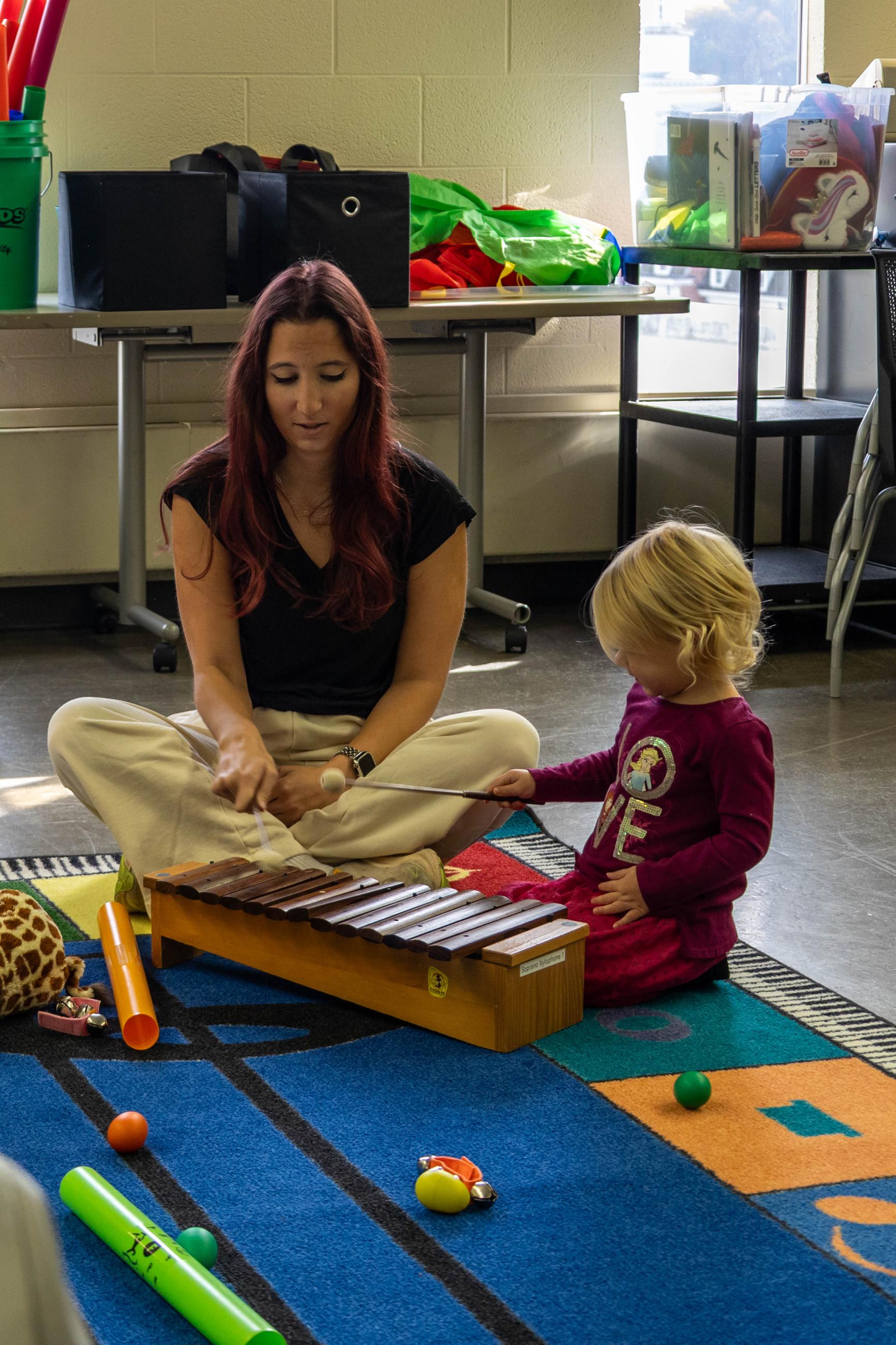A teacher sits cross-legged on a colorful rug with a toddler