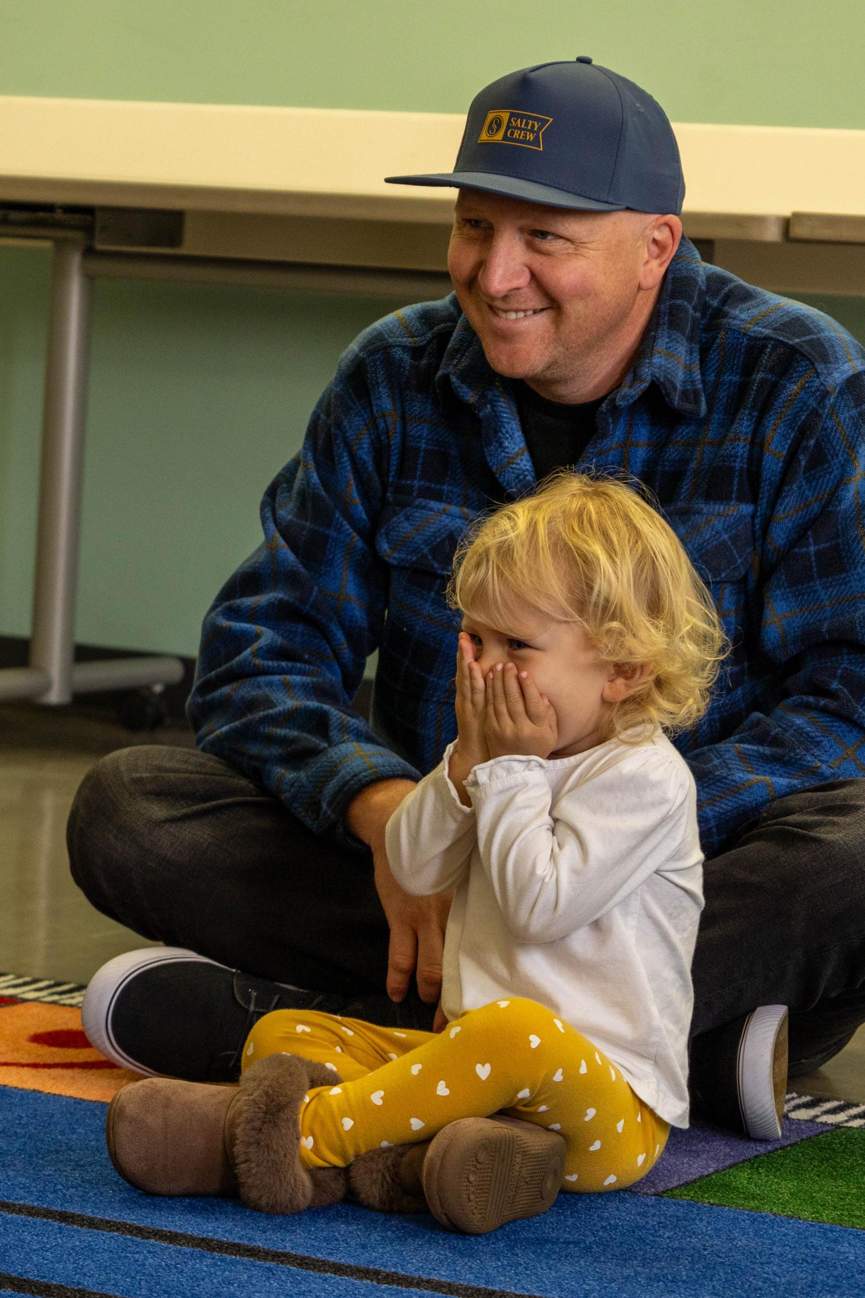 Father and giggling toddler sit on a classroom rug.