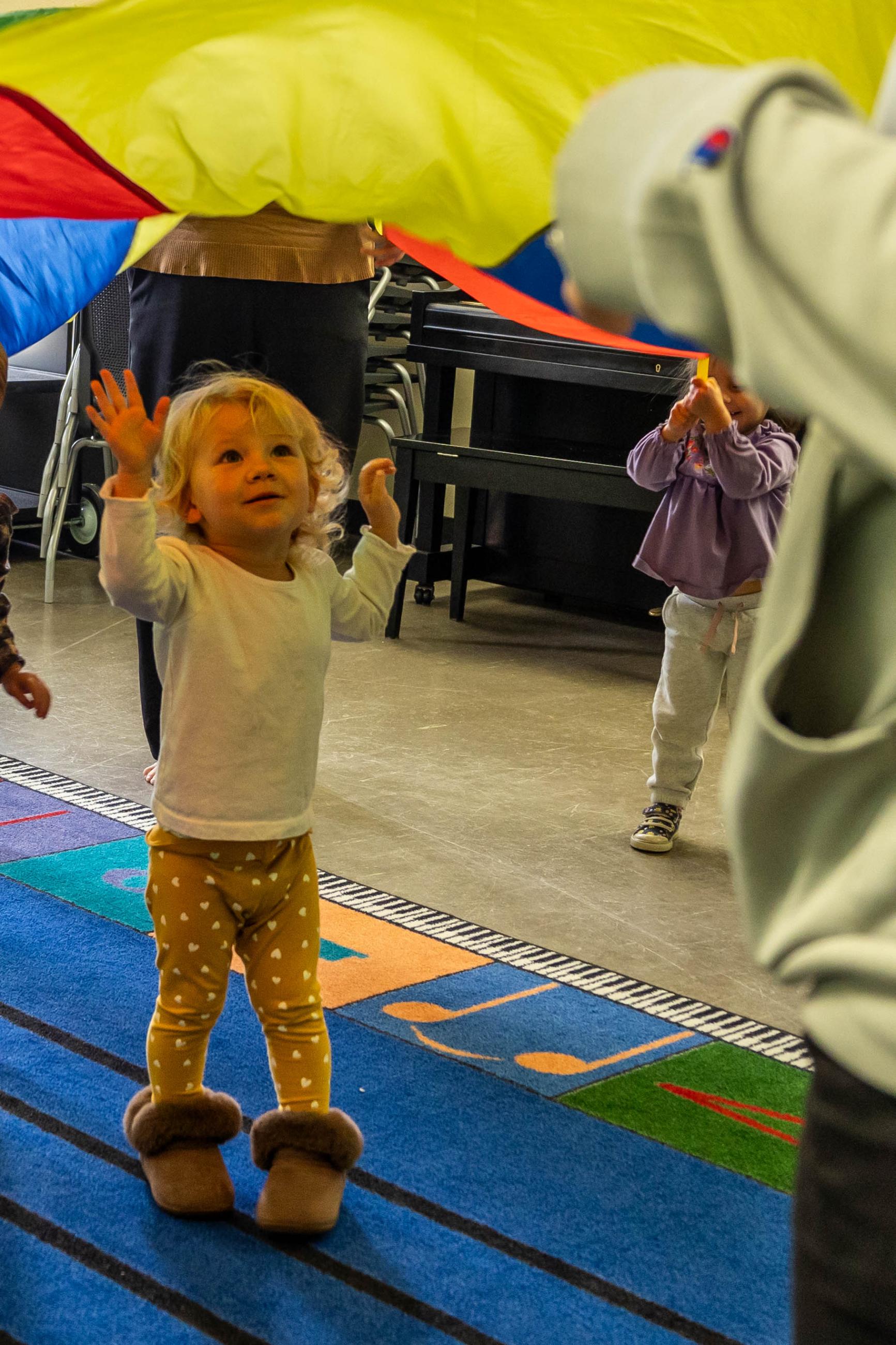 Toddler reaches up toward a rainbow parachute.