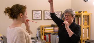 Two people in a room engaging in a vocal examination using red straws