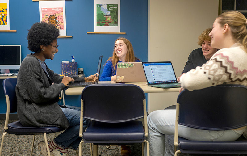 a group of students sitting at a table working on their computers while smiling and laughing