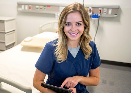 A woman wearing blue scrubs sits on a hospital bed while using an ipad