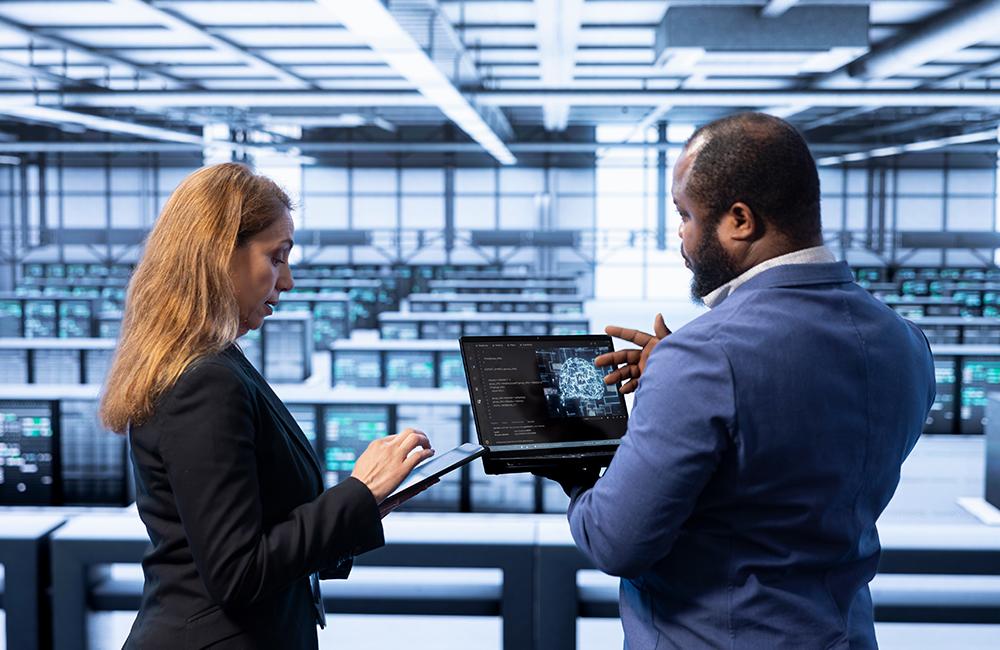 A man and a woman stand in a server room while working on a laptop and tablet