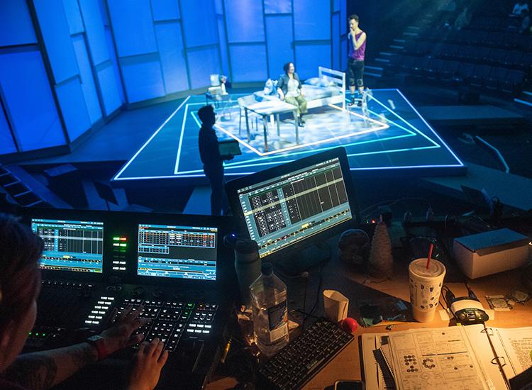 Students performing on a lit stage in the background while students in the foreground control the lighting and cameras from their computers