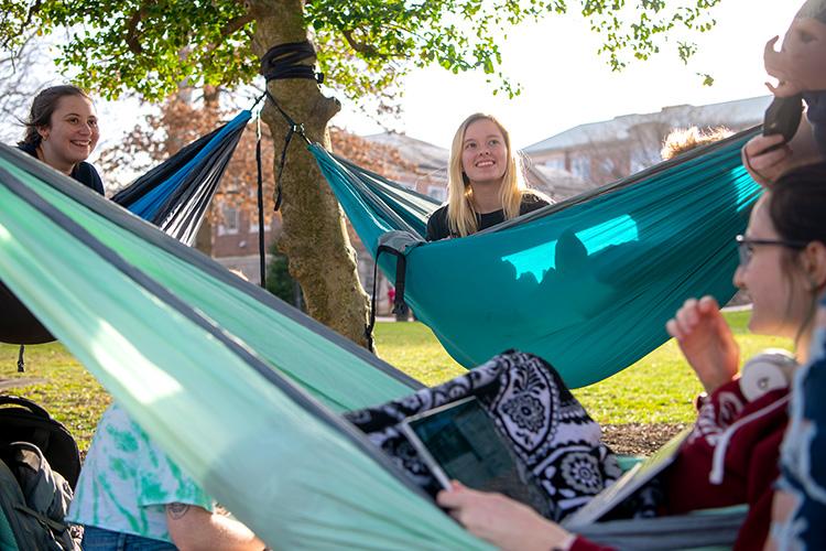students sit in hammocks that are hanging from trees on campus