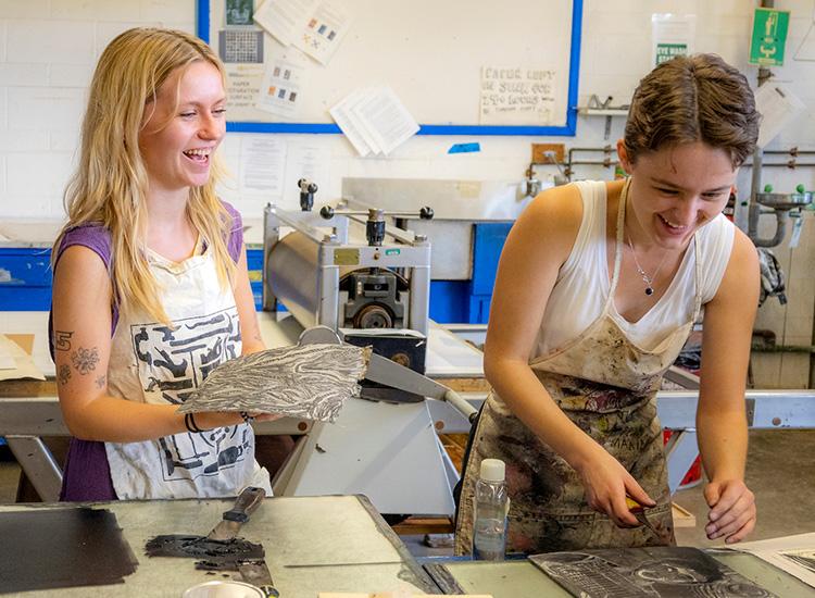 Two female students smile and laugh as they are working in the printmaking studio