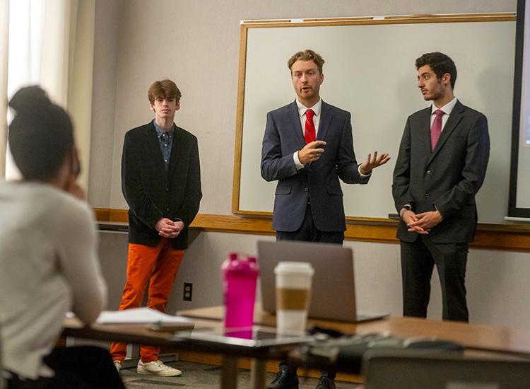 A group of male students wearing suits stand in front of a classroom and give a presentation