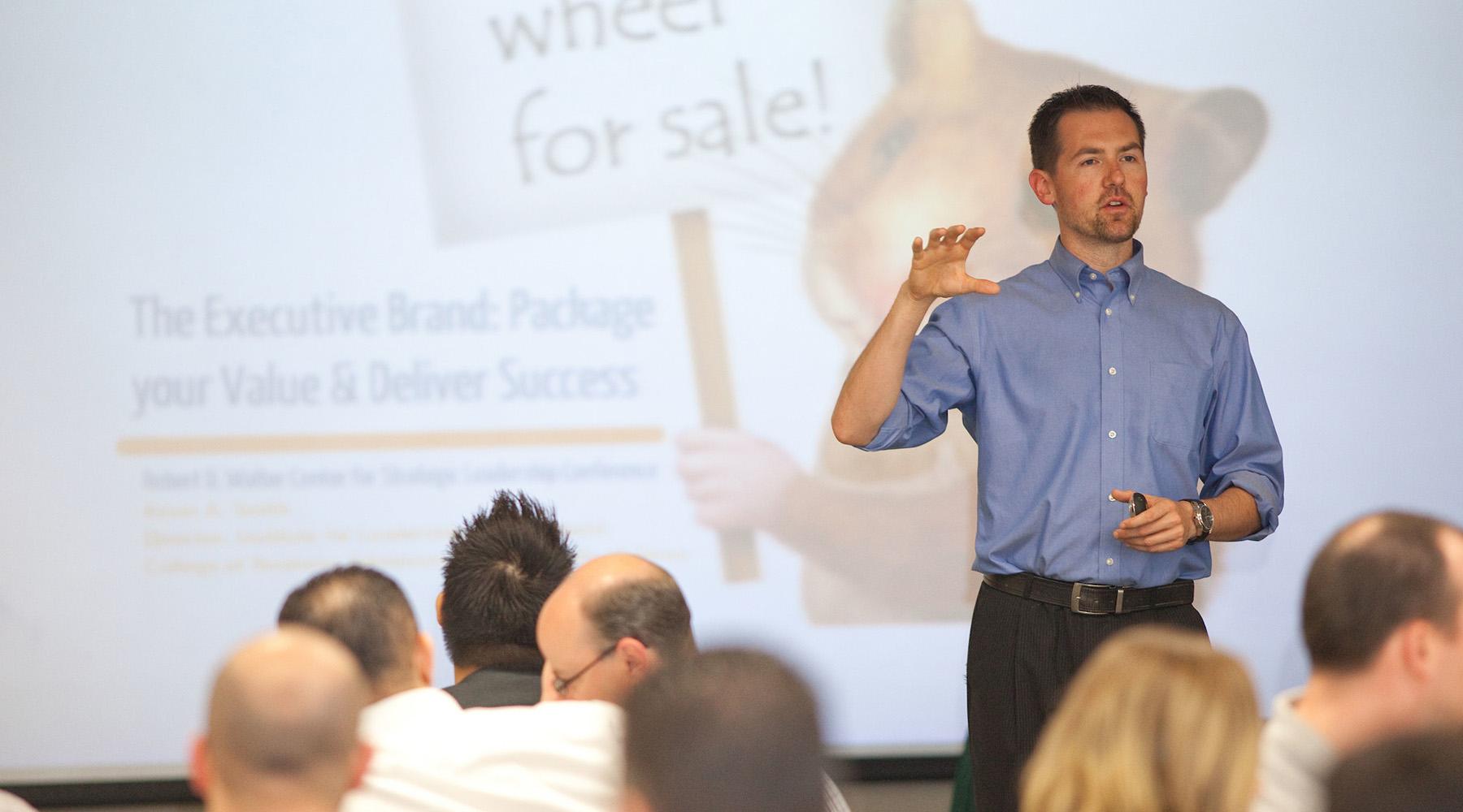 A man standing in front of a screen presents to a room full of people