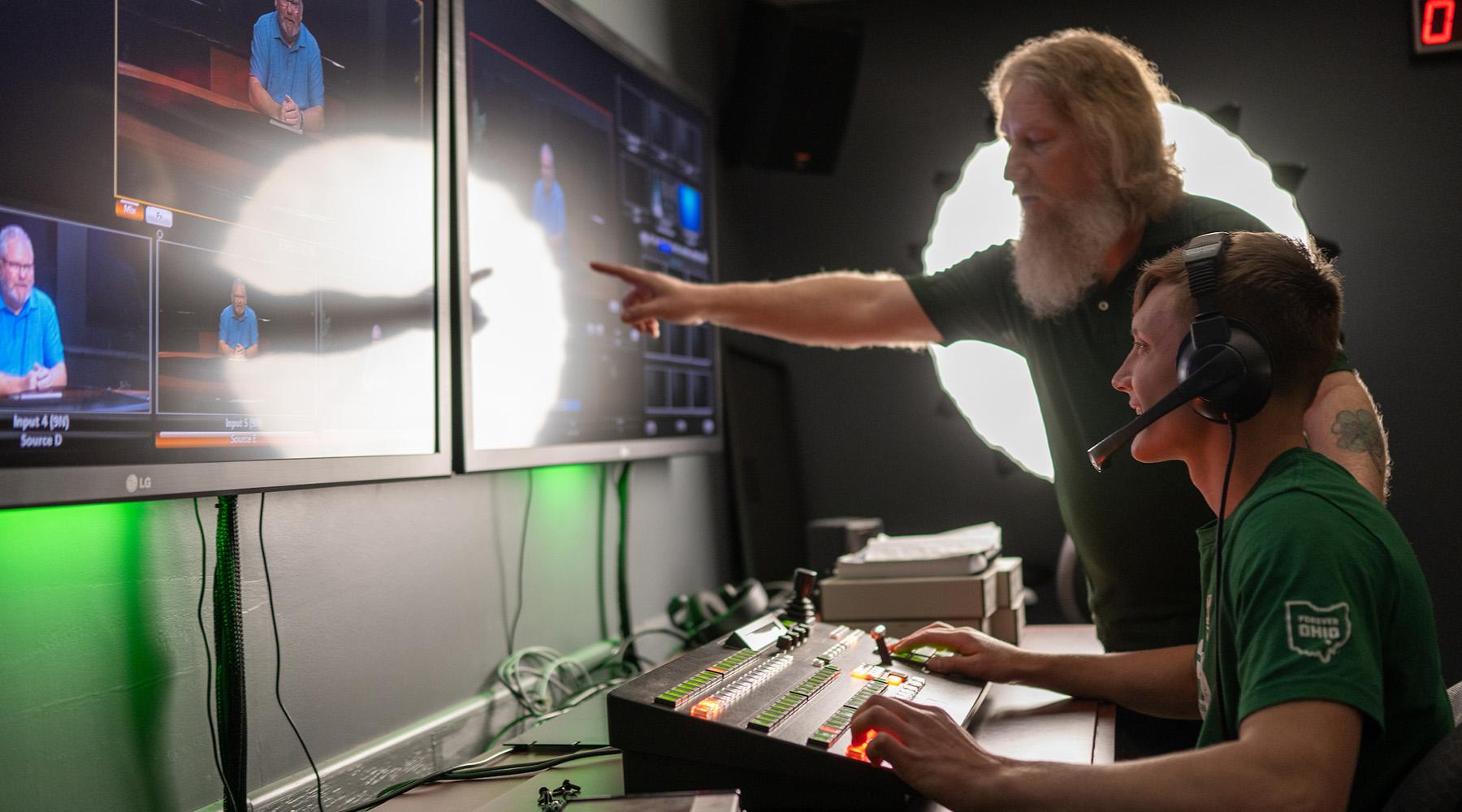 A professor points to a screen as a student sits at a control board while wearing a headset