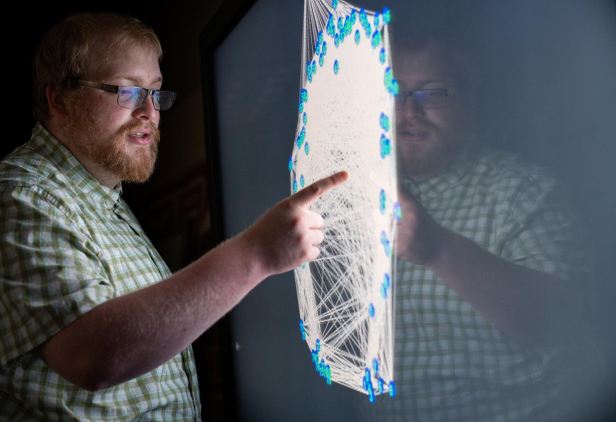 A man stands in front of and points to a screen displaying a network of colorful nodes