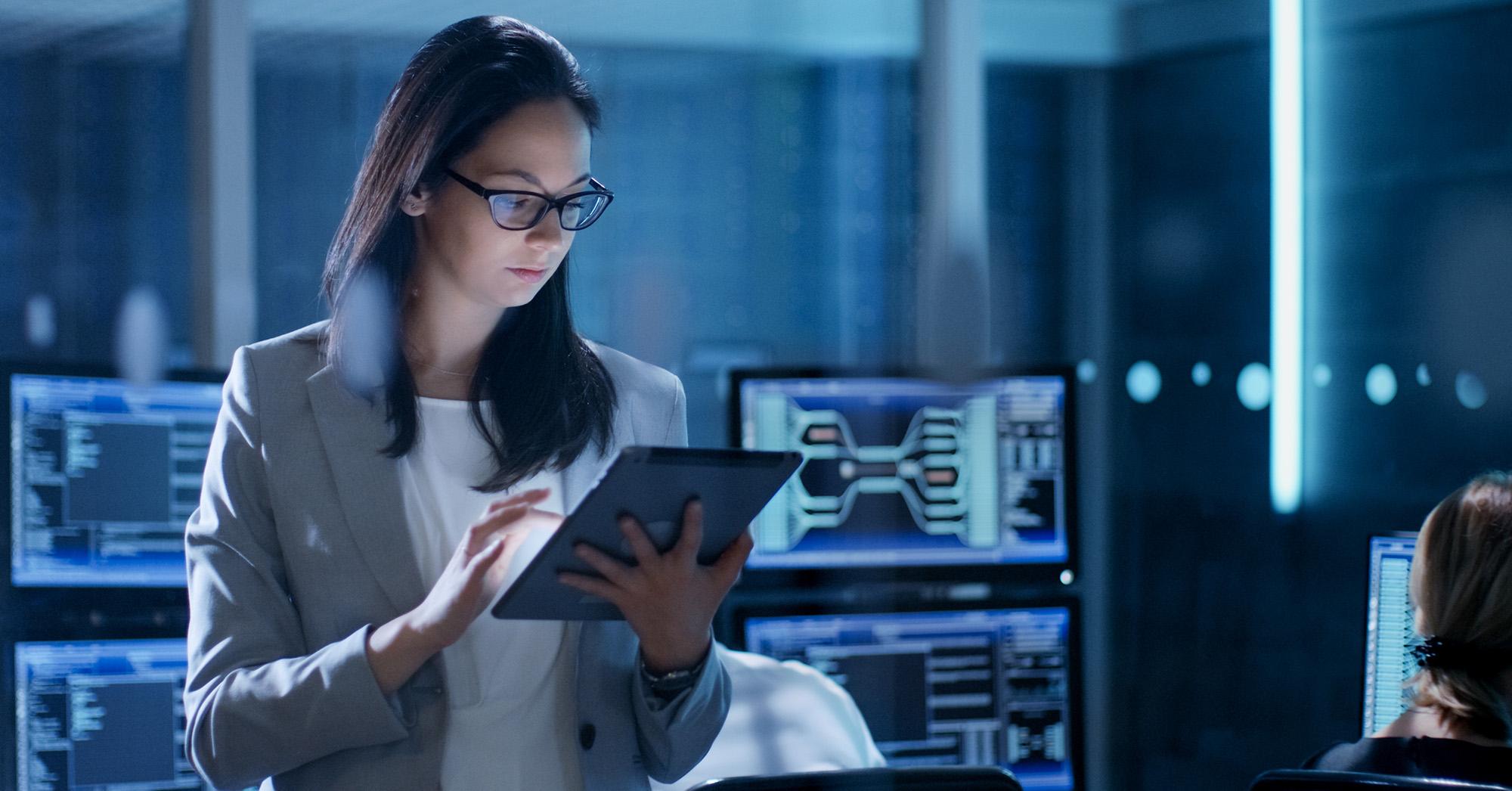 A woman using a tablet while standing in a room full of monitors
