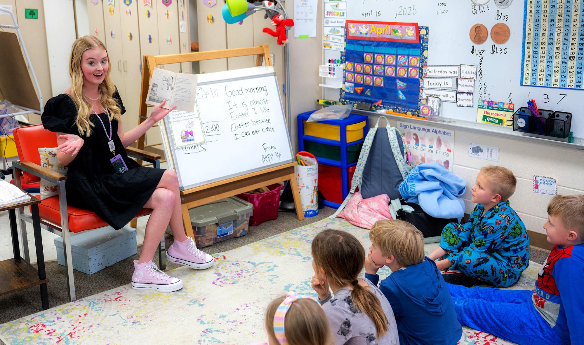 A female student teacher sits and reads a book to several elementary students