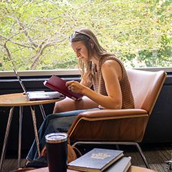 A female student sitting at a table reading a book and working on a laptop