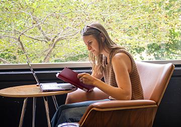 A woman sits at a small table reading a book with her laptop computer open on the table