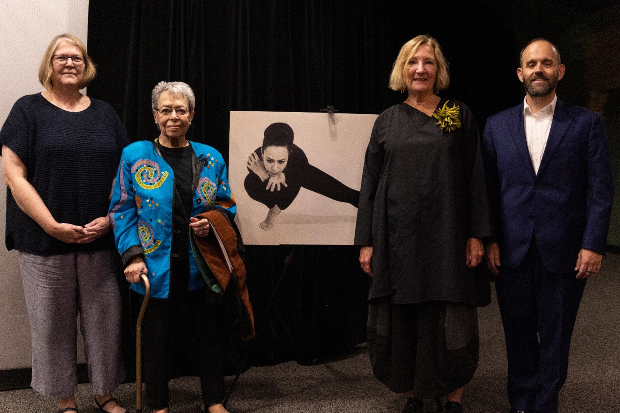 Picture of Glayds Bailin Stern, Elizabeth Sayrs, and Peggy Viehweger posing next to a large print of Gladys in a dance pose