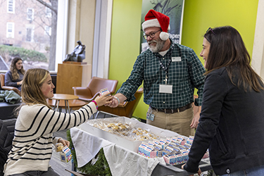 A man in a Santa Claus hat hands a packaged cookie to a student while a woman observes