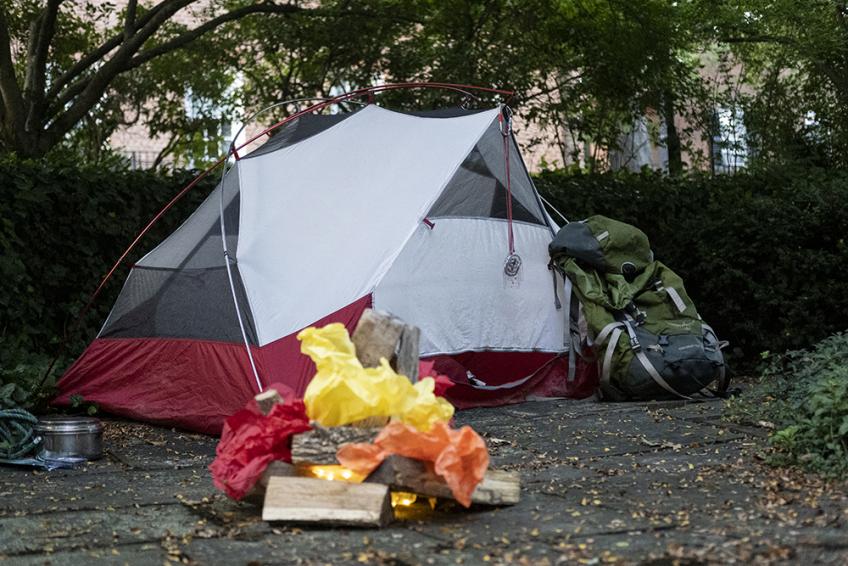 A small tent sits outside Alden Library, with a backpack leaning against it. Nearby is a simulated "campfire" with flames made of tissue paper.
