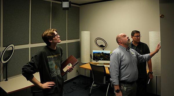 3 men stand in the Multimedia Studio, observing something on the wall above head level