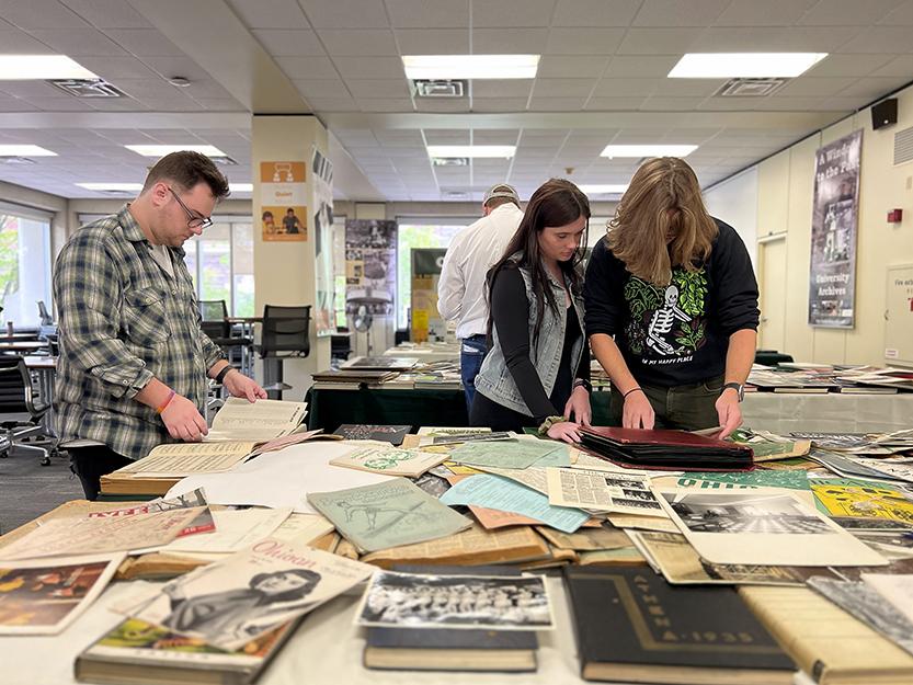Three college students peruse antique-looking books and magazines spread out on a table