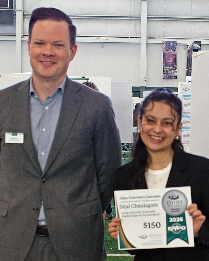 Person stands holding award certificate in front of their research poster. Tall man in grey suit stands next to them.