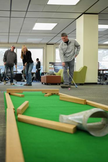 A man stands with one hand on his hip, attempting a one-handed golf putt in front of amused onlookers
