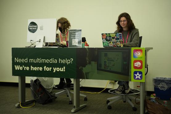 Two student employees are seated at the Multimedia Center help desk in Alden Library, prepared to assist patrons