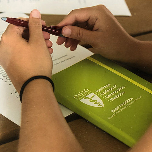 Close-up of a student's hands holding a pen next to an Ohio University Heritage College of Osteopathic Medicine pamphlet