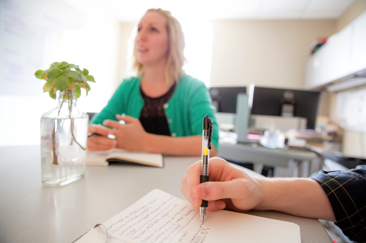 Two people sit at a table, one taking notes in a notebook.