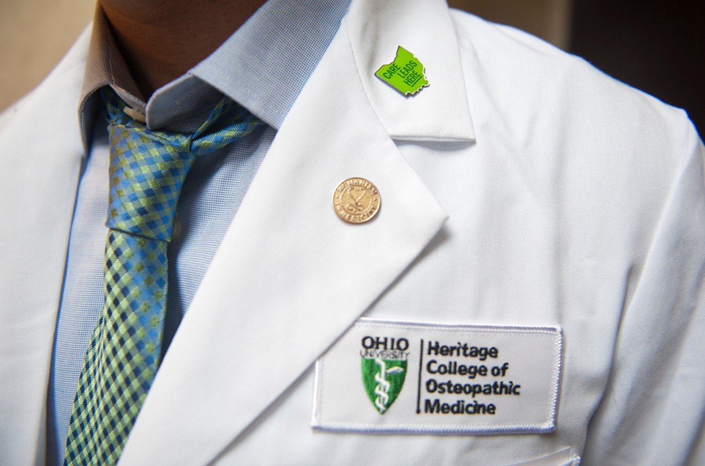 Person stands in white coat with HCOM logo and "Care Leads Here" pin.
