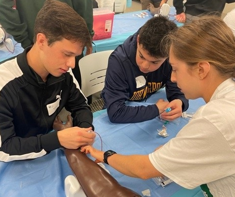 HCOM student assisting PreDOC participants with drawing blood from mannequin