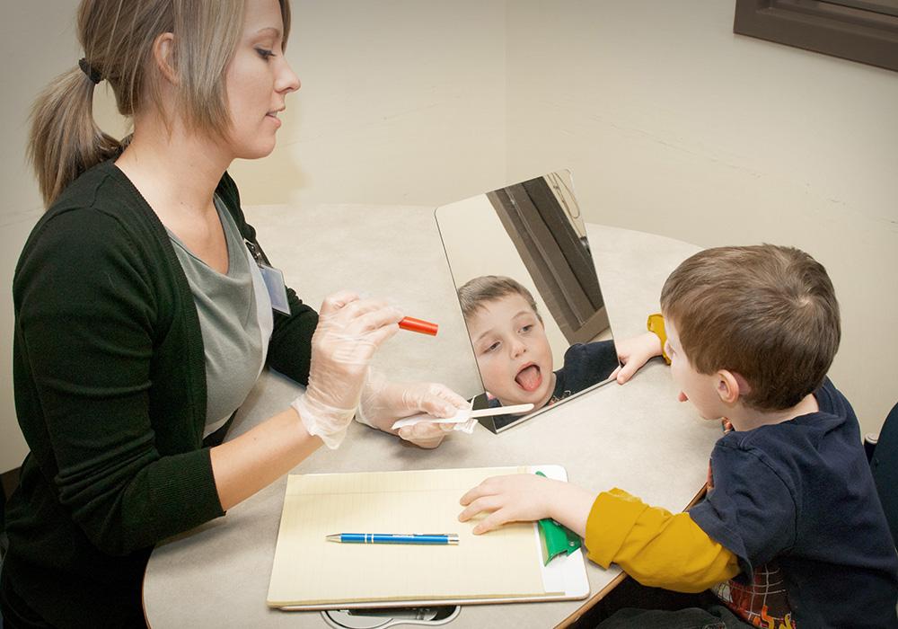 A woman sits at a table with a little boy as she tries to look into his mouth with a light and a tongue depressor