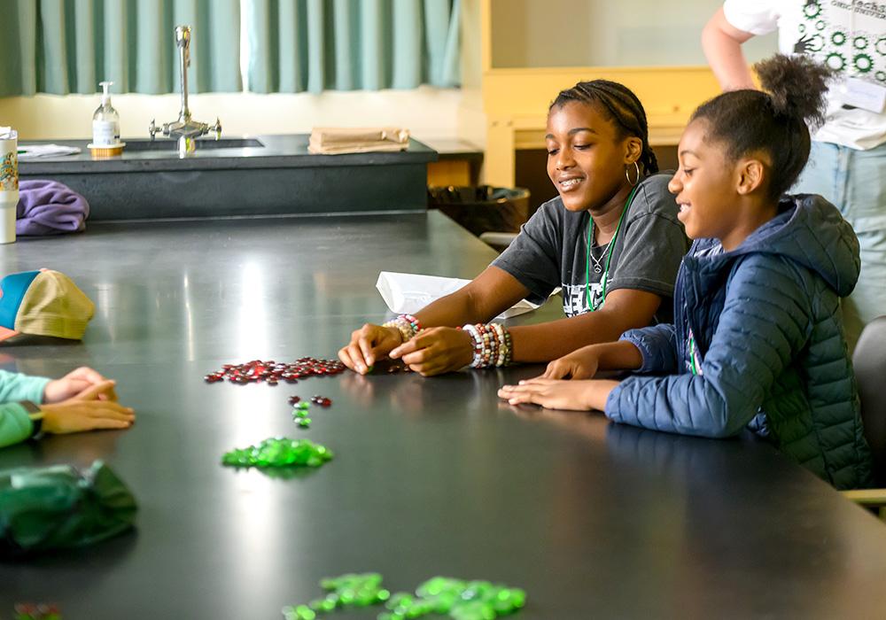 Two teenage girls sit at a table and work on a task using colored glass beads