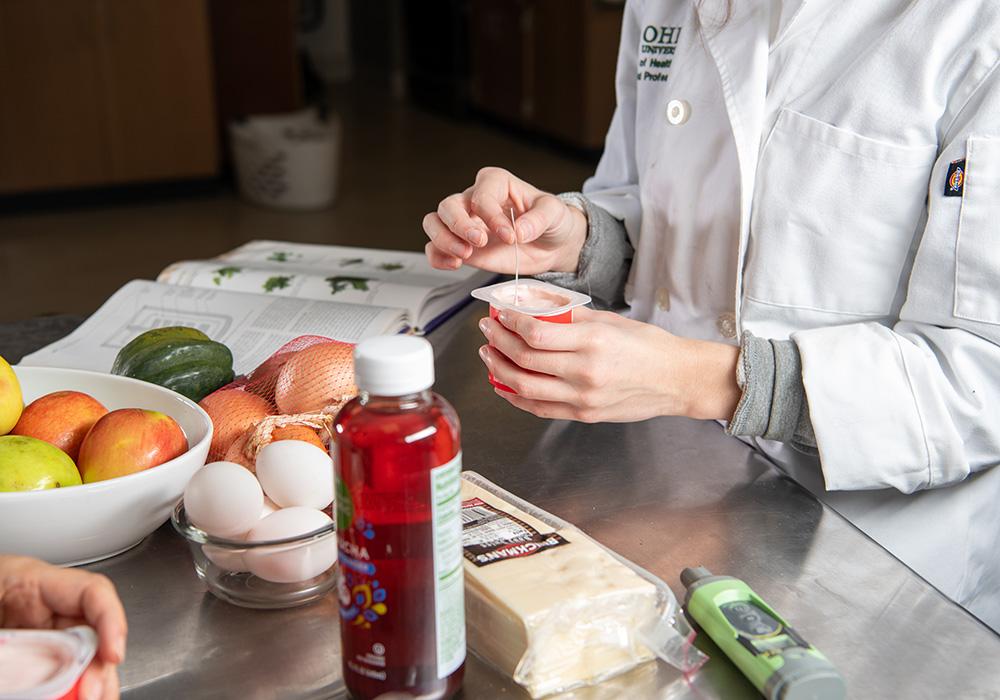 A woman holding an opened container of yogurt while standing at a table that is holding an assortment of other foods