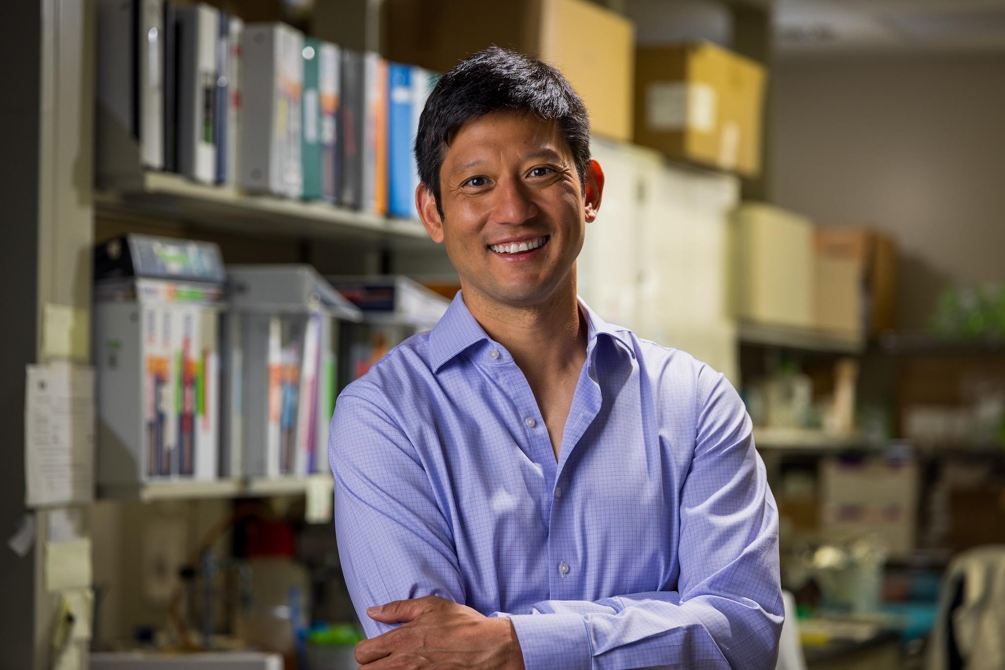 Dr. Kevin Lee in light blue dress shirt standing fron front of stacks of books and journals.