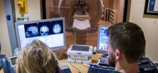 Two medical professionals look at MRI brain images on a computer while a patient lies in the machine in the background