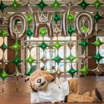OHIO's mascot, Rufus, poses for a photo in a white coat in front of balloons that spell out "OHIO."