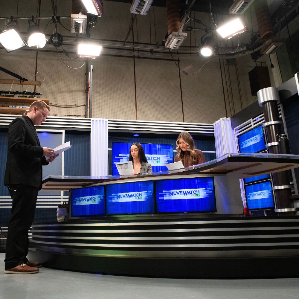 Two women sit at a news desk while a man stands in front of them