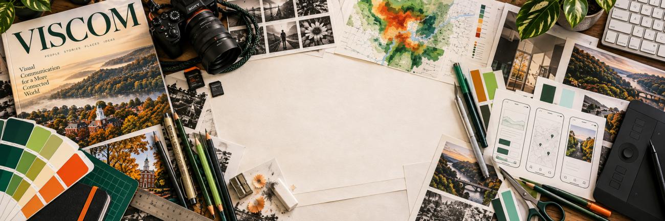 Overhead view of VisCom publications, campus photographs, infographics, and interface sketches layered with a camera, drawing tablet, pencils, and design tools around an open center
