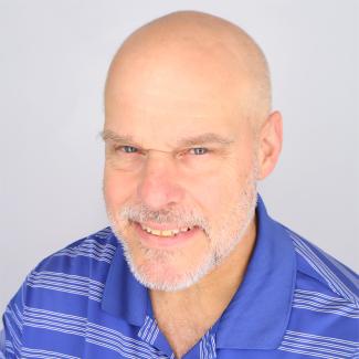 A person with a shaved head, grey beard and mustache and blue eyes wearing a blue polo with white stripes.