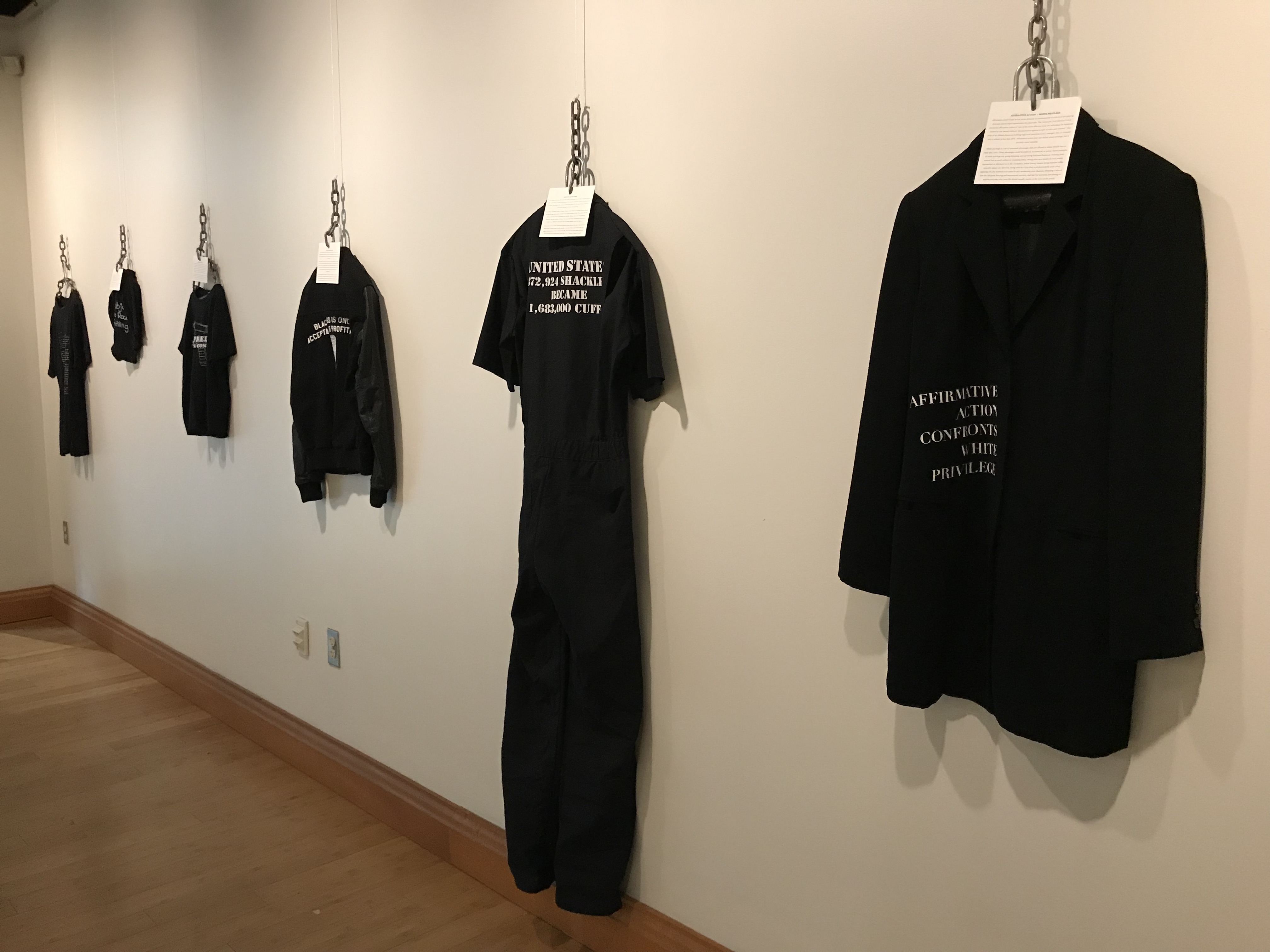 Various black clothing hung by chains in a row with bold white font printed on it with various statements about race