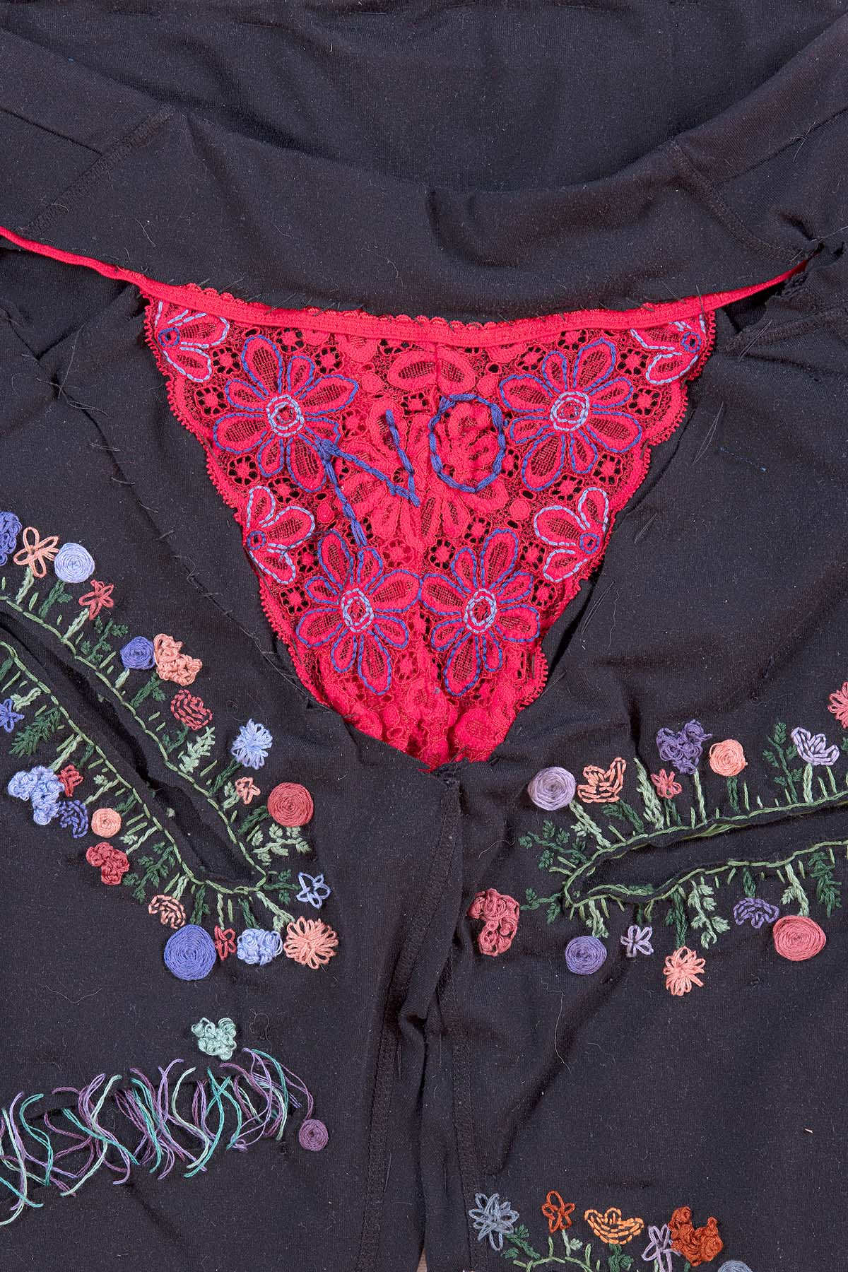 A close-up photograph of a red underwear garment visible atop black pants with the word 'NO' embroidered onto the garment