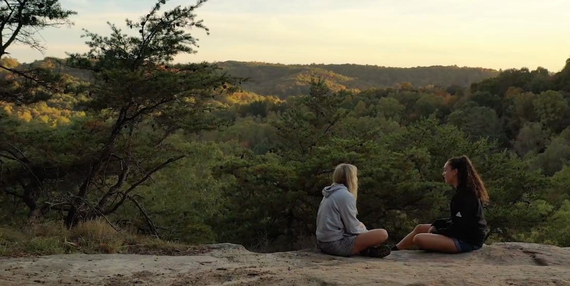two students sit on a rock in the forest after a hike